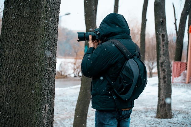 Un Homme Dans Une Veste Avec Une Capuche Et Une Mallette Sur Le Dos, Tenant Une Caméra Et Prenant Des Photos D'un Arbre