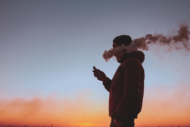 Un homme dans une veste fume une cigarette électronique à l'extérieur Vaping sur un fond de coucher de soleil