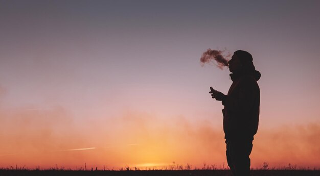 Un homme dans une veste fume une cigarette électronique à l'extérieur Vaping sur un fond de coucher de soleil