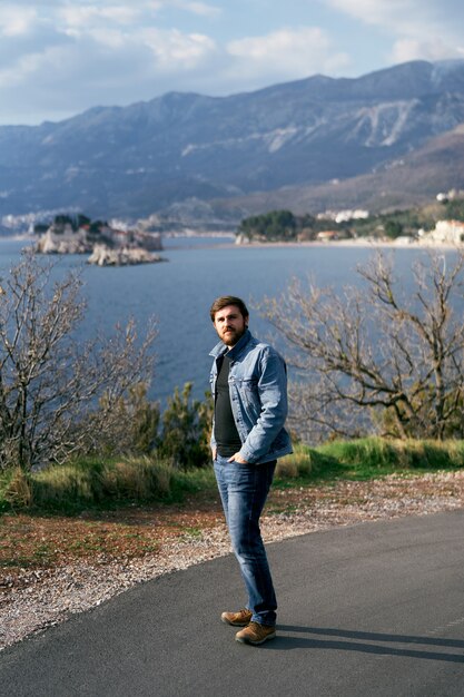 Homme dans une veste en jean bleu et un jean se tient en tenant ses mains dans ses poches sur une route goudronnée