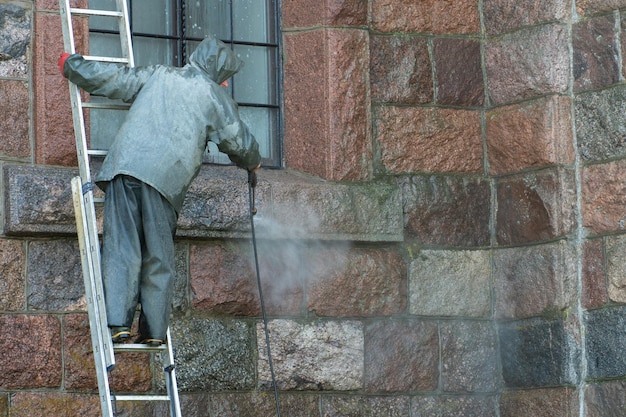 Un homme debout sur un escabeau lave les murs du bâtiment avec un jet d'eau sous pression Combinaison de protection chimique Décontamination du bâtiment du coronavirus Mesures de sécurité du covid19