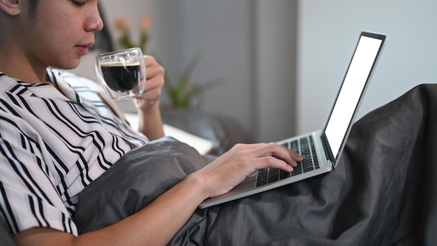 Homme décontracté buvant du café chaud et utilisant un ordinateur portable sur son lit le matin