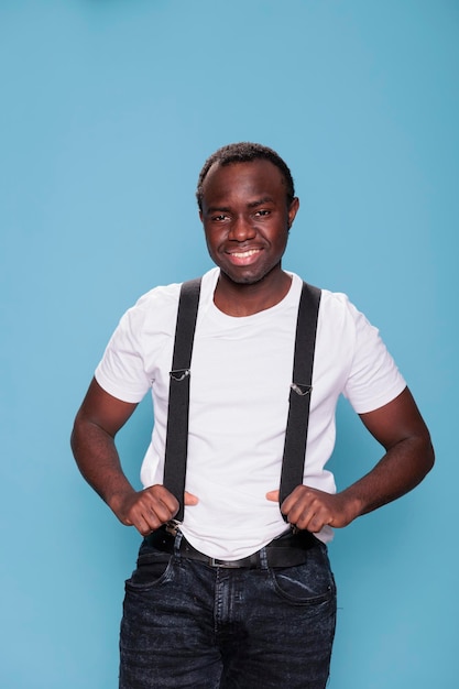 Homme décontracté posant pour la caméra en se tenant debout sur fond bleu. Souriant chaleureusement attrayant jeune homme adulte avec un style de mode portant un t-shirt blanc tout en tirant des bretelles noires.