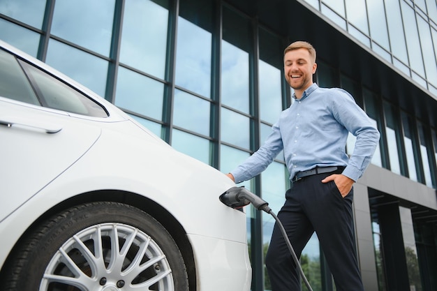 Homme décontracté avec smartphone près d'une voiture électrique en attente de la fin du processus de charge de la batterie