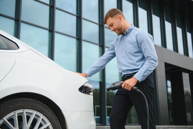 Homme décontracté avec smartphone près d'une voiture électrique en attente de la fin du processus de charge de la batterie