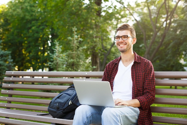 Homme décontracté souriant dans des verres travaillant avec un ordinateur dans le parc dans une atmosphère agréable. Concept de technologie, de communication, d'éducation et de travail à distance, espace de copie
