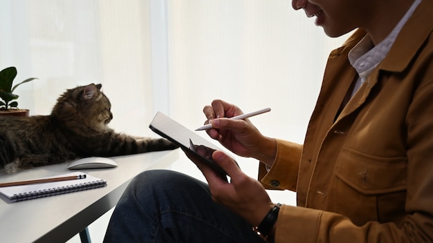 Photo homme décontracté tenant le câblage du stylo sur un ordinateur portable et assis avec un joli chat à la maison.