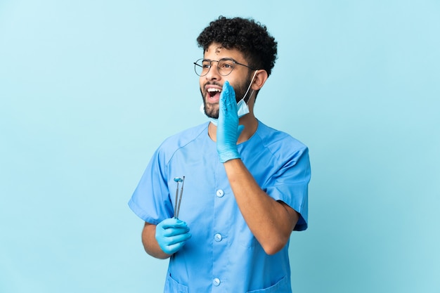 Homme dentiste marocain tenant des outils sur bleu criant avec la bouche grande ouverte sur le côté