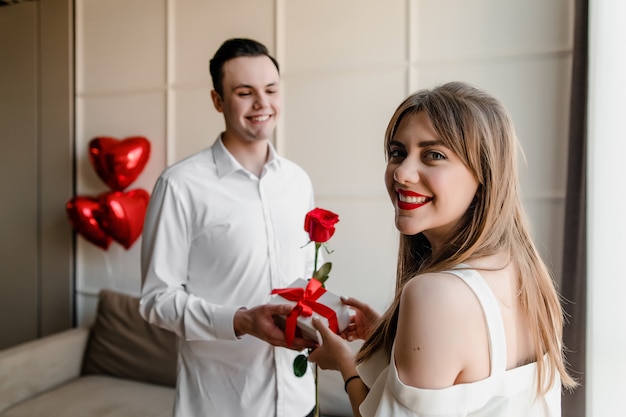 L'homme donne une rose avec un cadeau à la femme à la maison avec des ballons rouges en forme de coeur