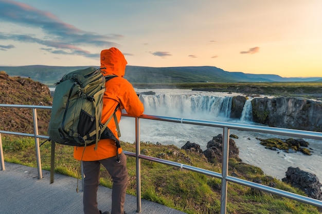 Homme à dos de sac à dos en veste orange debout et regardant la cascade de Godafoss au coucher du soleil pendant l'été en Islande