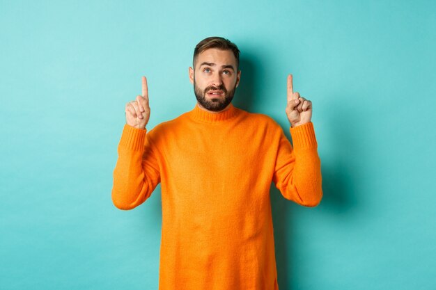 Homme douteux et non amusé à la recherche, pointant du doigt quelque chose de désagréable, debout sceptique sur fond turquoise.