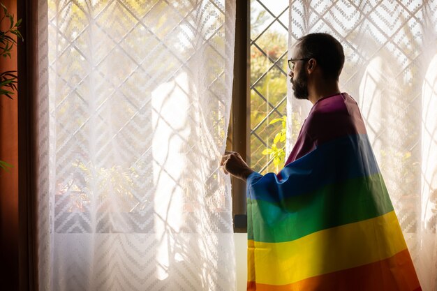 Homme avec drapeau lgbt sur les épaules en regardant par la fenêtre à l'extérieur