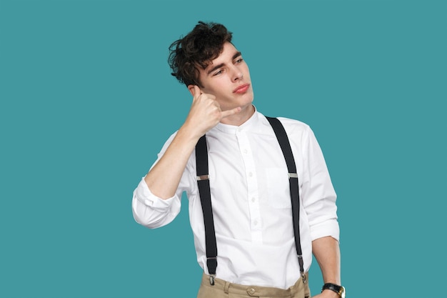 Homme drôle avec un geste d'appel sur les oreilles. Portrait de beau jeune homme d'affaires bouclé hipster en chemise blanche décontractée classique et jarretelle debout. studio intérieur tourné isolé sur fond bleu.