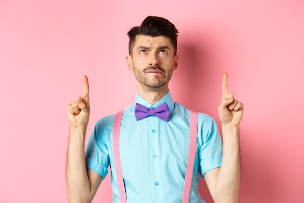 Homme drôle jaloux et triste en noeud papillon, mordant la lèvre et à la tentation de l'offre promotionnelle, pointant le doigt vers le haut, debout sur fond rose.