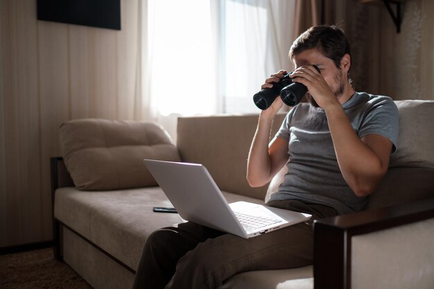 Homme drôle à la recherche de jumelles un ordinateur portable sur la table travaillant au bureau à domicile