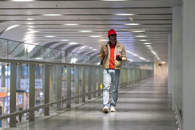 Homme du millénaire hipster noir marchant dans le terminal de l'aéroport à l'aide de téléphone portable en discutant dans les réseaux sociaux