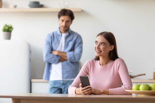 Un homme du millénaire triste et offensé regarde une femme souriante avec un smartphone dans la cuisine