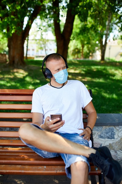 Homme écoutant de la musique avec un casque, à l'aide de smartphone portant un masque de protection du visage en plein air dans le parc