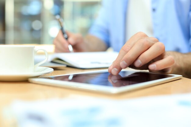 Homme écrivant un plan de travail avec un stylo sur un cahier en papier, naviguant sur Internet sur une tablette numérique sur un bureau.