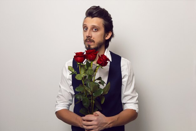 Homme élégant avec une barbe et des lunettes le jour de la Saint-Valentin dans une chemise blanche et un gilet sur un mur blanc se dresse avec une rose rouge