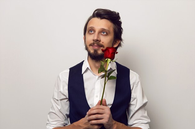 Homme élégant avec une barbe et des lunettes le jour de la Saint-Valentin dans une chemise blanche et un gilet sur un mur blanc se dresse avec une rose rouge