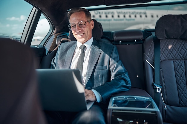 Un homme élégant et gai dans des verres est assis dans une voiture avec un ordinateur portable tout en étant transféré à l'aéroport