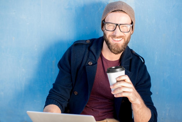Homme élégant habillé décontracté en pull et chapeau travaillant avec un ordinateur portable assis avec un café près du fond de mur bleu