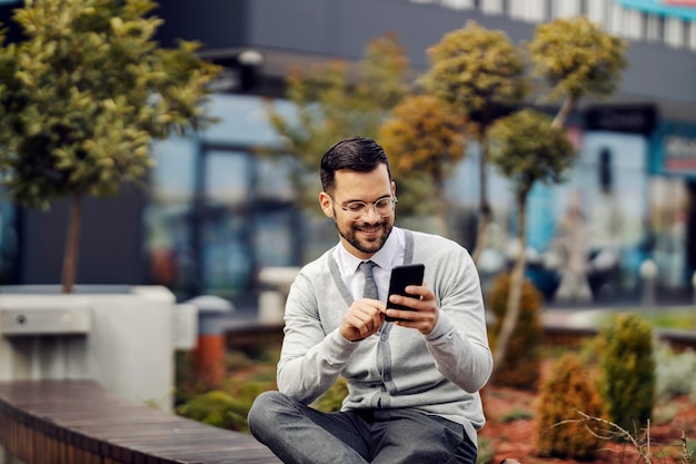 Un homme élégant et heureux est assis sur un banc dans le parc et tape au téléphone tout en souriant