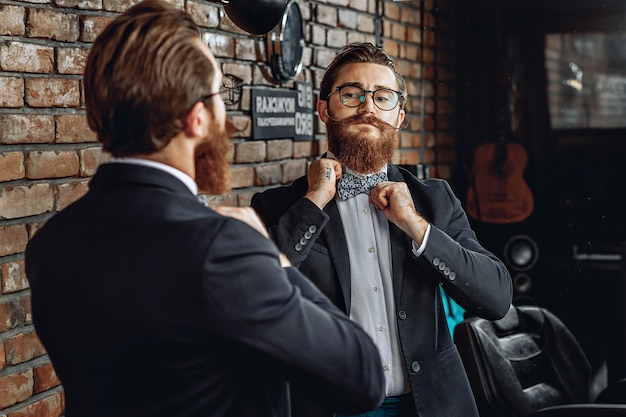 homme élégant avec des lunettes, une barbe et une moustache, se regardant dans le miroir et redressant un nœud papillon sur son col.