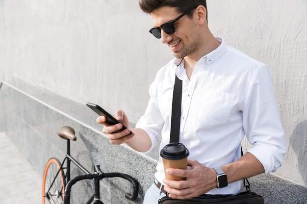 Homme élégant portant des lunettes de soleil, buvant du café à emporter et utilisant un téléphone portable en se tenant debout avec un vélo le long du mur extérieur