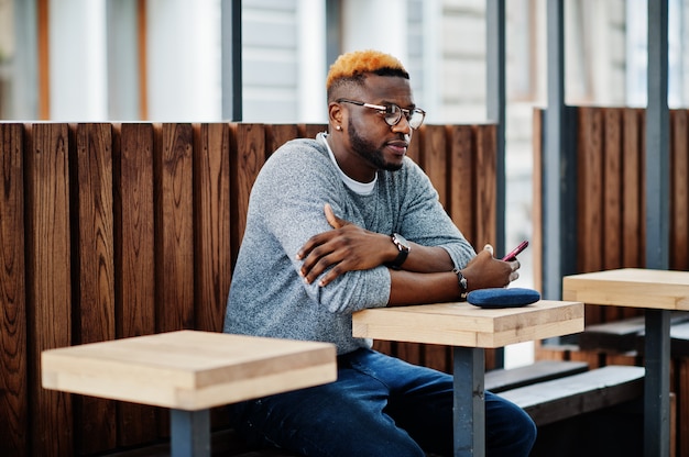 Homme élégant Sur Un Pull Gris Et Des Lunettes Posées Dans La Rue, Assis Sur Une Table, Café En Bois Avec Téléphone Portable