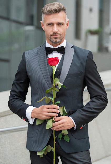 Homme élégant en smoking noir homme portant un smoking noeud papillon smoking grisonnant en plein air homme avec une rose rouge