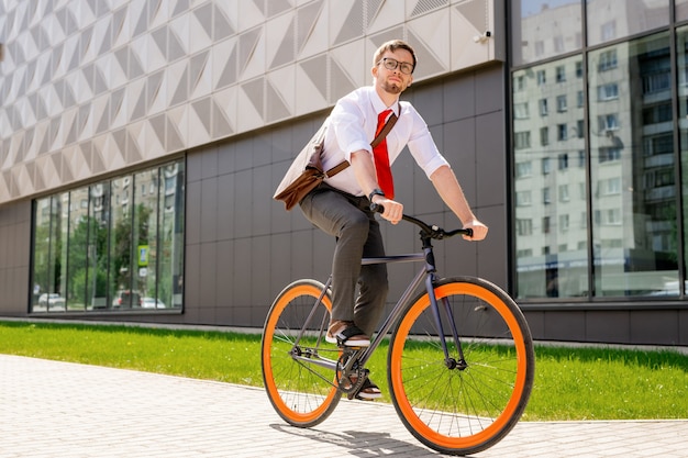 Homme élégant en tenue de soirée et lunettes à l'avant tout en allant à vélo en milieu urbain contre l'extérieur du centre d'affaires