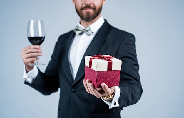 Homme élégant avec vue recadrée de visage non rasé en tenue de fête de vacances tenir un verre à vin rouge et une boîte-cadeau fond gris, célébration.