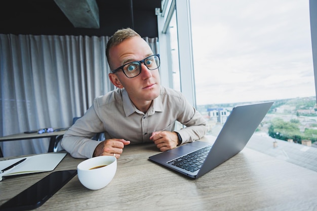 Homme émotif regardant la caméra sur le lieu de travail un homme à lunettes assis devant un bureau avec un ordinateur un étudiant cherche l'inspiration pour de nouvelles idées dans un café de bureau