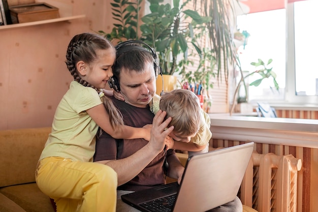 Homme avec enfants utilisant un ordinateur portable et des écouteurs pendant son travail à domicile, la vie en quarantaine