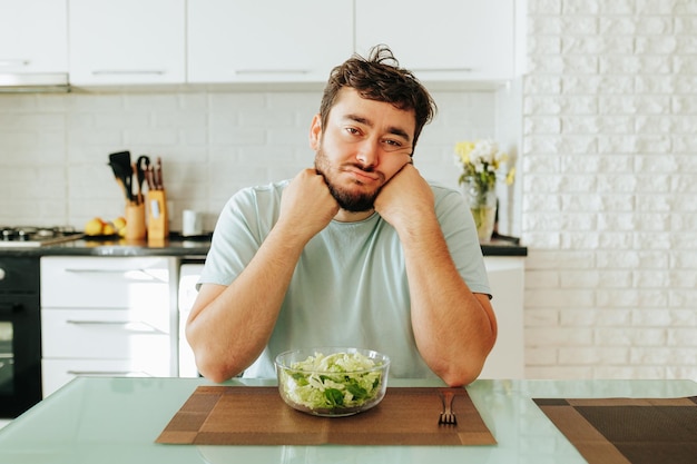 Un homme ennuyé est assis dans la cuisine avec sa tête dans ses mains devant une assiette de salade et une jeune fourchette