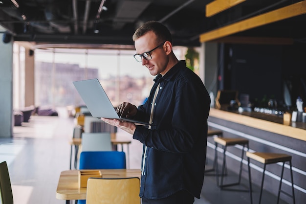 Homme entrepreneur en chemise noire et lunettes debout au bureau avec un ordinateur portable à la main patron d'entreprise prospère se sentant bien dans un style de vie riche