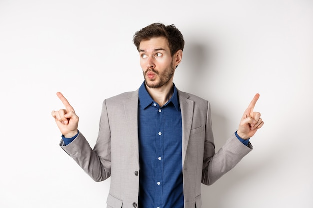 Homme entrepreneur indécis en costume pointant du doigt sur le côté, choisissant entre deux variantes, regardant le logo gauche pensif, debout sur fond blanc.