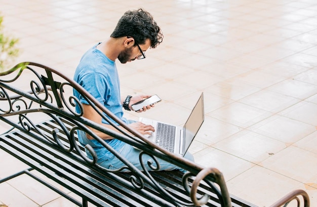 Homme entrepreneur travaillant avec un ordinateur portable et un téléphone portable dans un parc Homme indépendant travaillant avec un ordinateur portable tout en utilisant un téléphone jeune homme assis dans un parc travaillant avec son ordinateur portable et son téléphone portable