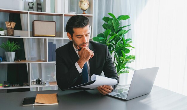 Un Homme Est Assis à Un Bureau Devant Un Ordinateur Portable Et Regarde Un Document.