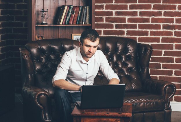 un homme est assis sur un canapé luxueux et travaille derrière un ordinateur portable dans son bureau sur le fond de la bibliothèque