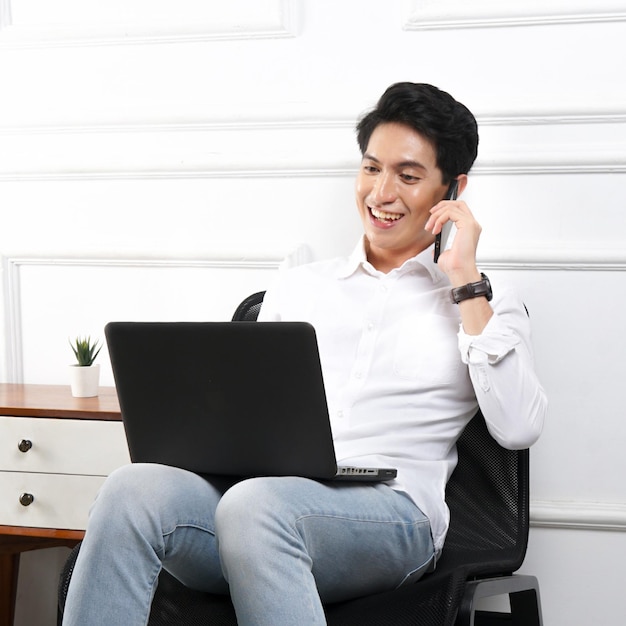 Un homme est assis sur une chaise avec un ordinateur portable et parle sur un téléphone portable.