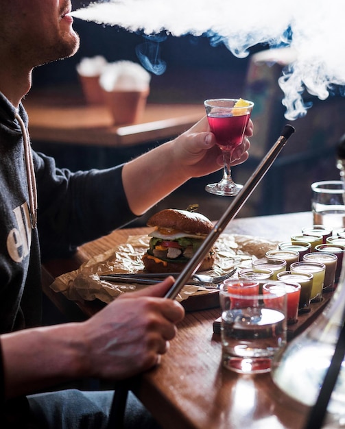 Un homme est assis dans un café boit de l'alcool mange un hamburger et fume un narguilé Photo verticale