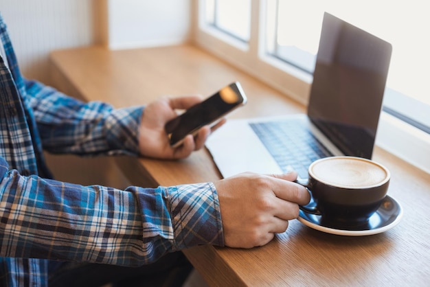 Un homme est assis dans un café et tient une tasse de café tout en travaillant sur un ordinateur portable et en utilisant un smartphone