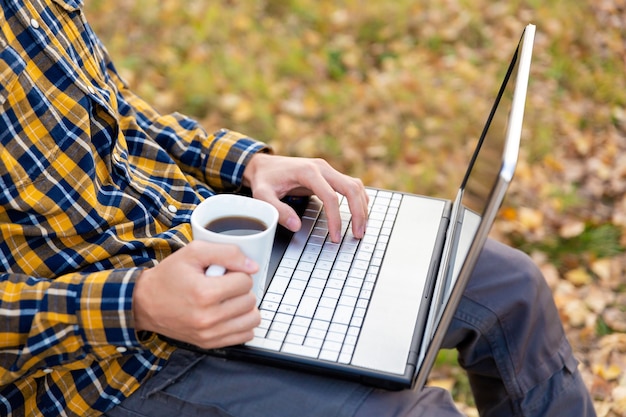 L'homme est assis dans le parc et travaille sur un ordinateur portable. Mains tapant sur le clavier et tenant une tasse de café