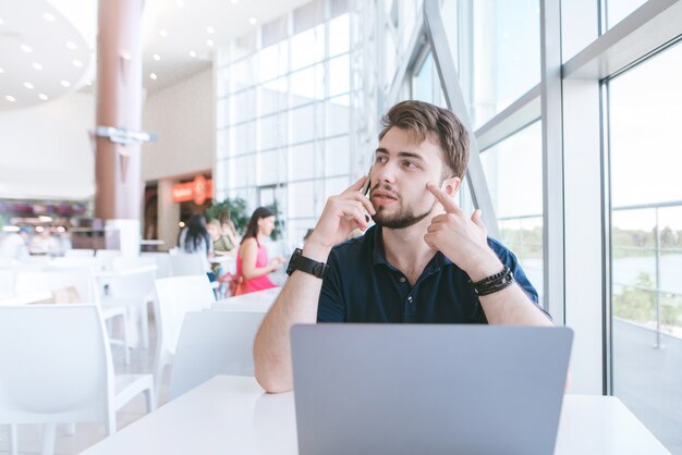 L'homme est assis dans un restaurant près de la fenêtre, travaille sur un ordinateur portable, parle au téléphone et regarde ailleurs.