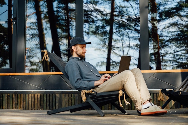 un homme est assis sur fond de fenêtres panoramiques d'une maison de campagne travaille dans un ordinateur portable