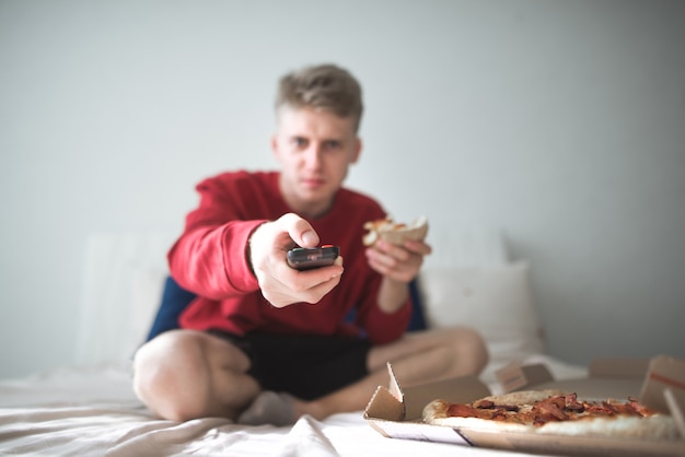 L'homme est assis à la maison dans un lit avec une boîte de pizza et montre à distance à huis clos