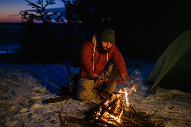 Un homme est assis près d'un feu de camp se réchauffant près du feu dans une forêt d'hiver au coucher du soleil